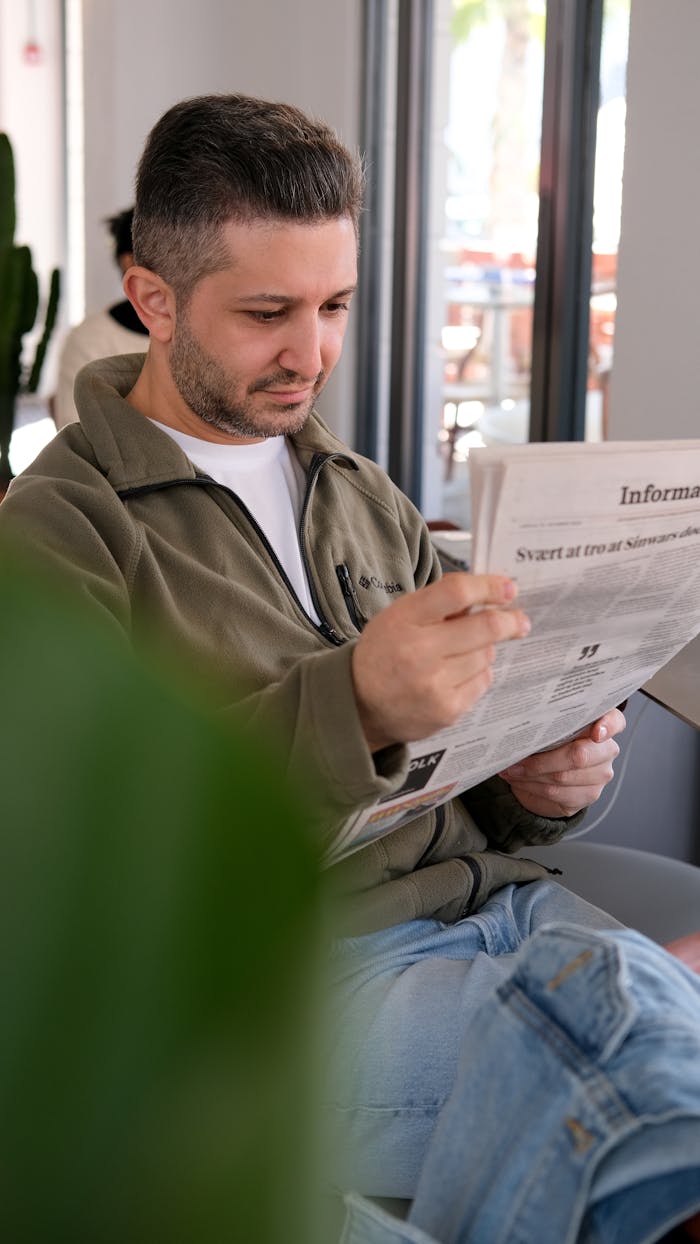 Man sitting in a cafe reading a newspaper in a relaxed setting. Daylight streams through windows.